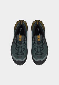 Ζευγάρι αθλητικών παπουτσιών σε σκούρο πράσινο και μαύρο με κίτρινες λεπτομέρειες, ελαστικό σύστημα κορδονιών και μαξιλαράκια εσωτερικά, πάνω σε απλό λευκό φόντο.