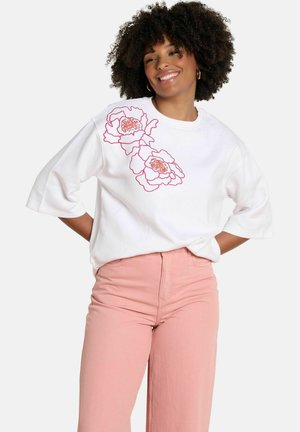 Uśmiechnięta kobieta o kręconych włosach, ubrana w biały top z różowymi kwiatowymi wzorami i różowe spodnie z wysokim stanem, dłonie na biodrach.