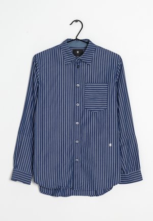 Camisa azul marino de manga larga con botones, con rayas blancas verticales y un bolsillo en el pecho a rayas, colgada en una percha negra.
