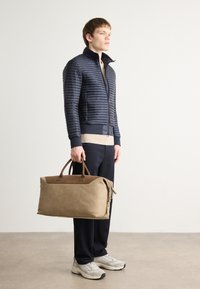 Veste bomber matelassée bleu marine avec des poignets côtelés, pull beige, pantalon foncé et un sac de voyage en cuir tan avec des accents en suède et des poignées doubles.