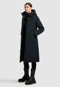 Femeie îmbrăcată într-o haină lungă de iarnă umplută cu puf, de culoare neagră, cu glugă, leggings negri și cizme negre cu șireturi, stând în fața unui fundal simplu și luminos.