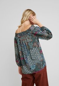 Poisin oksalle ulottuva pusero tummasta kuvioituneesta kankaasta, jossa on monivärisiä yksityiskohtia, frillaneck ja pallomaiset hihat.