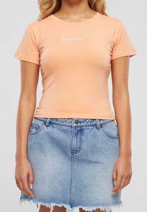 Femme portant un crop top ajusté à manches courtes couleur pêche et une jupe en jean bleu clair effilochée sur un fond uni.
