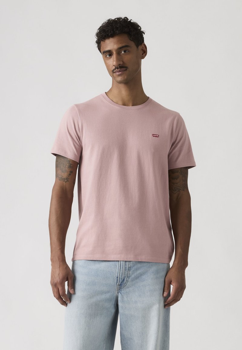 Homme aux cheveux courts bouclés et tatouages, portant un t-shirt Levi's rose clair et un jean bleu clair, debout devant un fond uni.