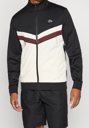 Homme portant une veste de sport zippée noire, blanche et bordeaux avec un petit logo de crocodile vert sur le côté gauche de la poitrine et un short noir.