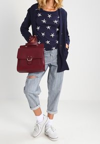 Burgundi nahka käsilaukku, jossa on rakenteellinen muotoilu ja yläkahva. Mallilla on yllään sininen tähti-kuvioinen paita, ylisuuria raidallisia farkkuja ja valkoisia tennareita.