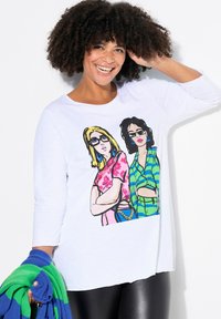 Maglietta bianca a maniche lunghe con una grafica colorata di due donne in outfit vivaci. La texture appare morbida; include un accessorio lavorato a maglia blu e verde.