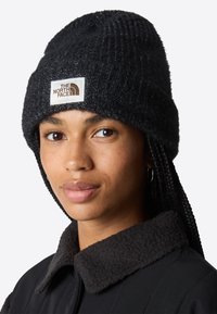 Berretto in maglia nero con una trama a costine e un'etichetta con logo. Presenta alette per le orecchie intrecciate ed è abbinato a una giacca nera con colletto in pile.
