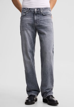 Person trägt locker sitzende, ausgewaschene graue Jeans, schwarze Lederschuhe und ein in die Hose gestecktes weißes Hemd, steht vor einem schlichten hellen Hintergrund.