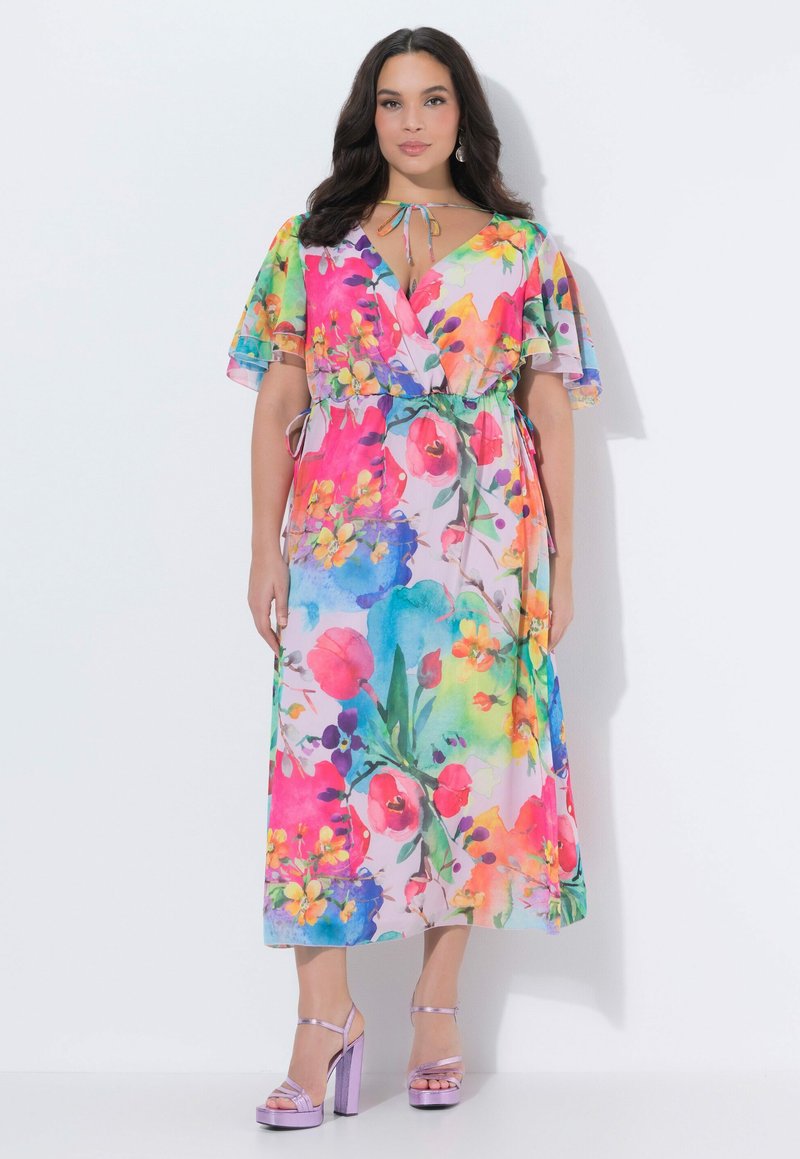 Žena v barevných květovaných zavinovacích šatech s volánkovými rukávy a vázáním na krku, doplněných lesklými levandulovými sandály na vysokém podpatku.