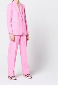Completo rosa con giacca aderente e pantaloni a gamba larga, caratterizzato da un tessuto texturizzato e un delicato motivo a pois, abbinato a sneaker colorate.