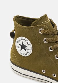 Olive green suede high-top sneaker featuring a black star logo patch, silver eyelets, and white rubber sole with black stripe.