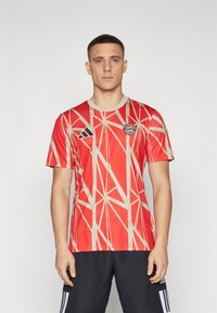 Camiseta deportiva roja con patrón geométrico en marrón claro, con escote redondo y mangas cortas. Logo en el pecho y marca en el hombro.
