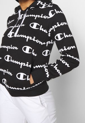 Sweat à capuche noir avec motif texte du logo Champion en blanc, cordons blancs, porté avec un pantalon blanc, main de la personne dans la poche.