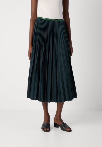 Jupe midi plissée bleu canard avec une ceinture verte ornée d'un logo ; associée à des mules ouvertes noires. Le tissu a une texture lisse, offrant une silhouette fluide.