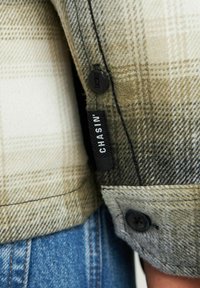 Camicia a quadri in toni beige e oliva, con una morbida texture in lana, bottoni neri e un'etichetta rettangolare con il logo "CHASIN'".