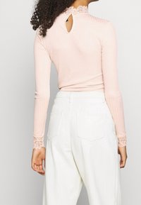 Ljust rosa ribbat långärmad topp med spetsdetaljer vid kragen och ärmsluten, med en nyckelhålsöppning i ryggen. Matchad med vita byxor.