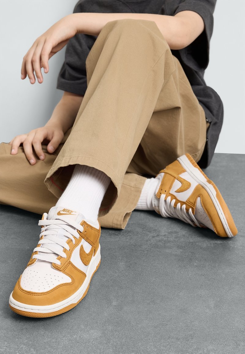 Personne assise par terre portant un pantalon beige, des chaussettes blanches et des baskets Nike blanches et orange avec les lacets attachés.