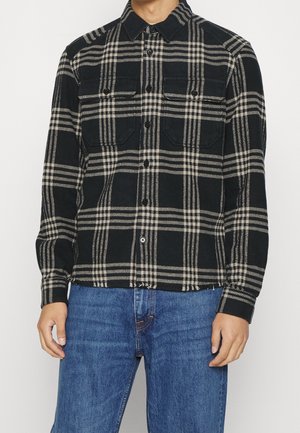 Uomo che indossa una camicia a quadri nera e beige con tasche sul petto e jeans blu, in piedi con le braccia rilassate.