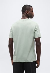 Άνδρας, με την πλάτη γυρισμένη, φοράει ανοιχτό πράσινο κοντομάνικο πουκάμισο και μαύρο παντελόνι μπροστά σε απλό λευκό φόντο.