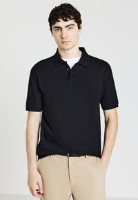 Polo shirt nera a maniche corte, tessuto strutturato, colletto con bottoni, abbinata a pantaloni beige chiaro. Design pulito e moderno.