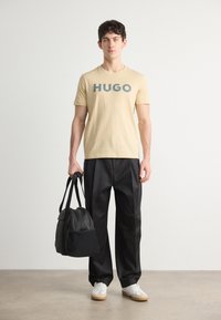 Bēša kokvilnas T-kreklu ar tumsni pelēku uzdrukātu "HUGO". Melni bikses un melna auduma soma ar plecu siksnām. Baltas kedas ar brūnām akcentiem.
