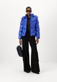 Jachetă puffer albastră cu un finisaj lucios, glugă și închidere cu fermoar. Purtați-o peste un top negru transparent și asociați-o cu pantaloni negri largi și o geantă.