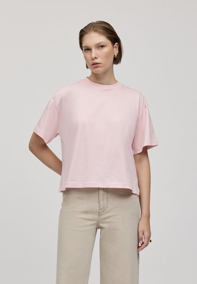 T-shirt raccourci rose clair, à manches courtes, col rond, en coton doux, porté avec un pantalon beige taille haute. Conception simple et minimaliste.