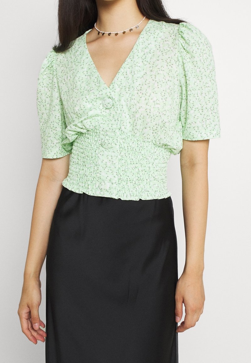 Blusa floreale verde chiaro con maniche a sbuffo corte, vita smock e dettagli con bottoni; abbinata a una gonna nera. Tessuto dalla texture morbida.