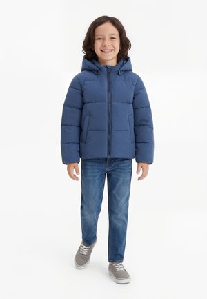 Gyerek mosolyog, kék kapucnis steppelt dzsekit, kék farmert és szürke tornacipőt visel, kamerába sétálva, fehér háttér előtt.