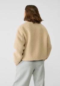 Beżowa kurtka z polaru o teksturowanej powierzchni i krótkim fasonie. Posiada wysoki kołnierz i luźne rękawy, zestawiona z szarymi spodniami.