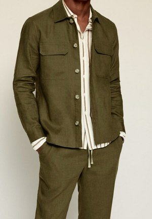 Mann trägt eine olivgrüne Jacke mit Knöpfen und passende Hose, darunter ein weiß-beige gestreiftes Hemd, die Hände in den Taschen.