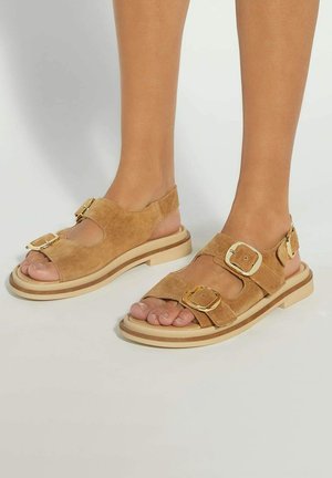 Tan suede sandals featuring two wide straps, gold buckles, and a layered sole with a light, chunky design. Open-toe with a back strap.