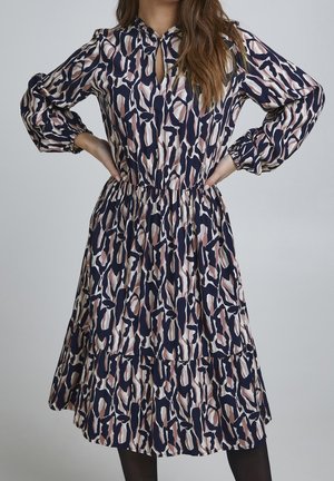 Femme portant une robe à manches longues, longueur genoux, avec des motifs abstraits bleu marine, beige et rose, debout les mains sur les hanches devant un fond gris.