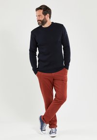 Marineblauer Pullover mit Rundhalsausschnitt und drei Knopfakzenten an der Schulter, kombiniert mit roten Hosen und marineblauen Sneakers mit weißen Schnürsenkeln.