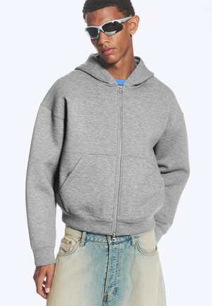 Joven con gafas de sol deportivas blancas, sudadera con capucha gris con cremallera y vaqueros azules desgastados, de pie frente a un fondo liso.