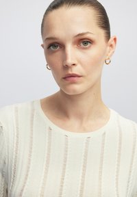 Heller cremefarbener Pullover mit vertikalem Streifenmuster und strukturiertem Strick. Runder Ausschnitt und dezente Rippung. Akzentuierung durch goldene Creolen.