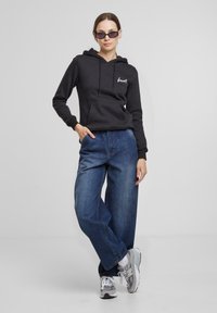 Čierna mikina s kapucňou a predným vreckom, s bielym logom, skombinovaná s voľnými modrými džínsami a šedými teniskami New Balance. Minimalistický dizajn.