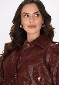 Hnědá kožená bunda s lesklým povrchem, vybavená límcem, zlatě tónovanými detaily a náprsními kapsami. Hladká textura s jemným vzorem.