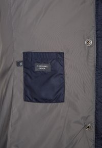 Giacca grigia con una tasca blu navy contenente un'etichetta. Il materiale è liscio con una leggera texture trapuntata. Include bottoni a pressione e un'etichetta sul lato.