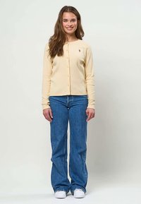 Jeune femme aux longs cheveux bruns portant un cardigan beige boutonné, un jean bleu et des baskets blanches, debout et souriante devant un fond uni.