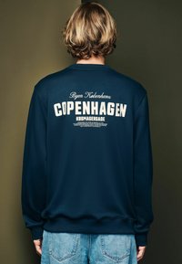 Námornícky modrý mikina s dlhými rukávmi, na chrbte s bielym textom "COPENHAGEN" a "Byen København". Mäkká tkanina a rebrované manžety.