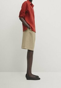 Chemise rouge en lin à boutons, short beige mi-long, chaussures plates tissées noires, texture lisse, manches retroussées, vue latérale de la tenue.