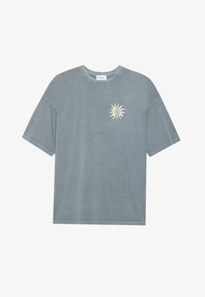 Gaiši zils, pārāk liels T-krekls, kas izgatavots no mīkstas kokvilnas, ar apaļu kakla izgriezumu un dzeltenu saules grafiku uz kreisā krūtis.