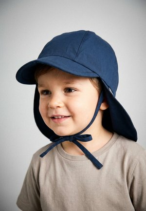 Young child wearing a beige shirt and a navy blue sun hat with chin straps tied in a bow, smiling and looking slightly to the side.