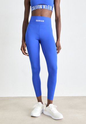Personne portant des leggings Calvin Klein bleu vif et un soutien-gorge de sport assorti, associés à des baskets blanches, se tenant sur un sol clair.