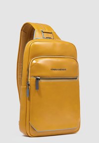 Bolso bandolera de cuero amarillo con textura suave, asa superior, bolsillo delantero con cremallera y costuras en contraste. Presenta un logo en la parte frontal.