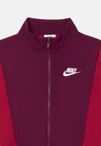 Bordo i crvena jakna s patentnim zatvaračem i visokim ovratnikom, s bijelim Nike logom na lijevoj strani prsiju i glatkom, laganom tkaninom.