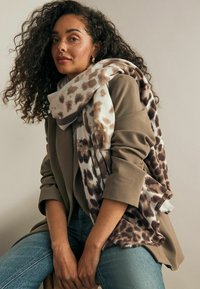 Een bruin-creme dierenprint sjaal hangt over een beige blazer, met franjes, gecombineerd met blauwe jeans in een zittende pose.