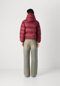 Giubbotto imbottito bordeaux con cappuccio, caratterizzato da un design trapuntato. Abbinato a jeans a gamba larga blu chiaro, che mostrano una vestibilità casual e rilassata.